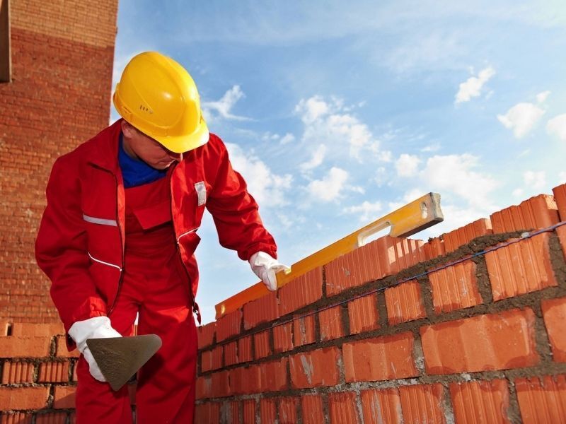 Obrero de la construcción con uniforme rojo y casco amarillo colocando ladrillos en una pared de ladrillos al aire libre.