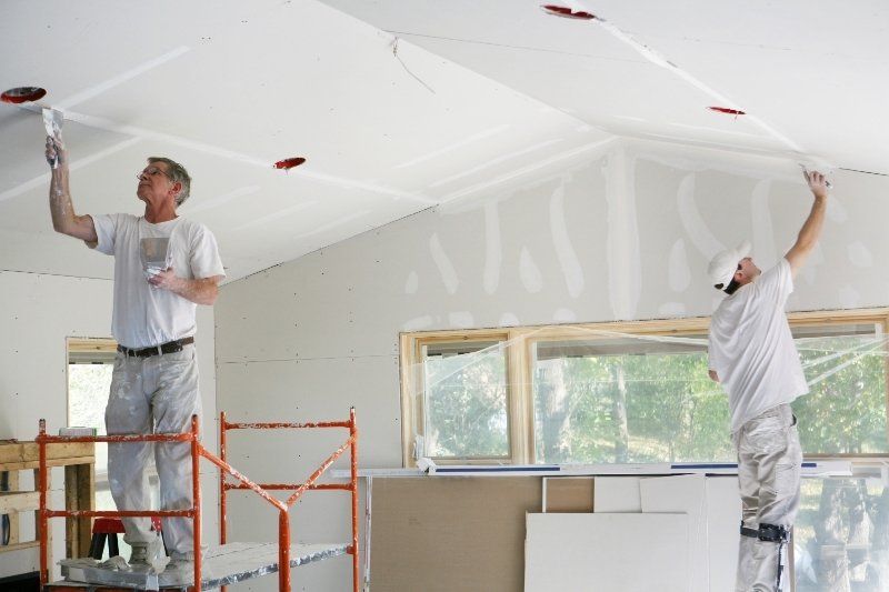 Dos trabajadores pintando un techo blanco en una habitación con andamios y grandes ventanales.