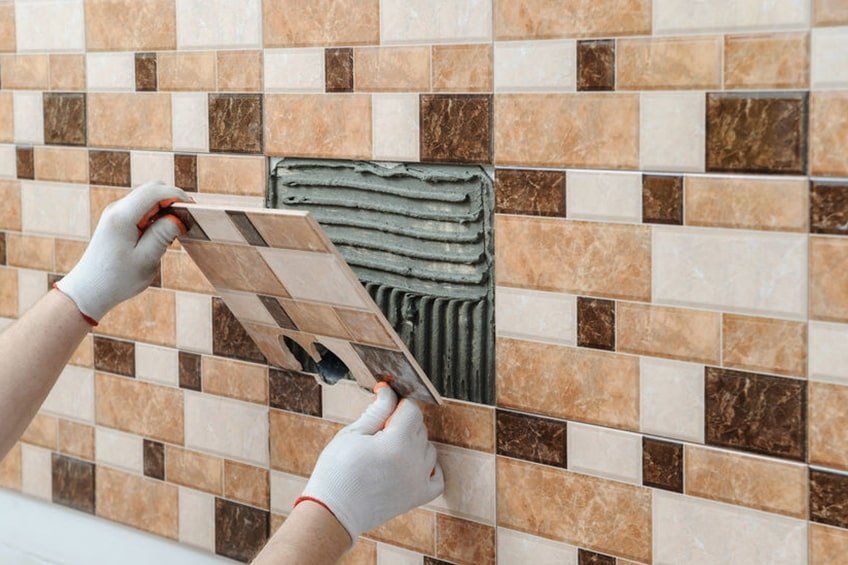 Manos colocando una baldosa estampada sobre una pared alicatada con adhesivo fresco, reparando un área de salpicadero.