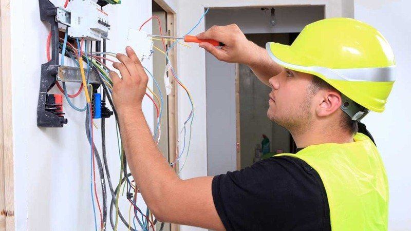 Electricista con casco amarillo cableando un panel eléctrico con herramientas en una abertura de la pared.