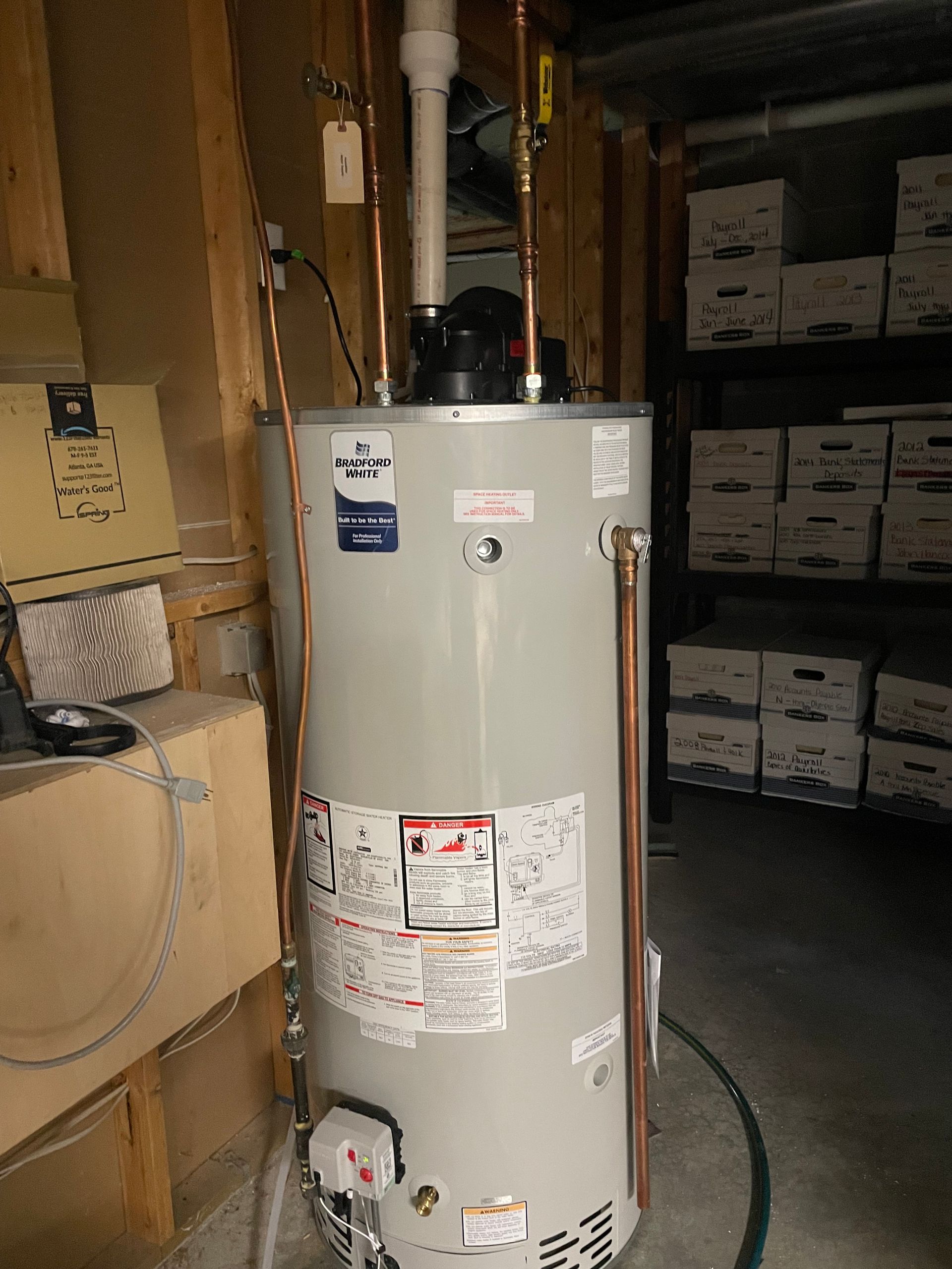 A tall, cylindrical water heater in a storage area, with copper pipes and ventilation.