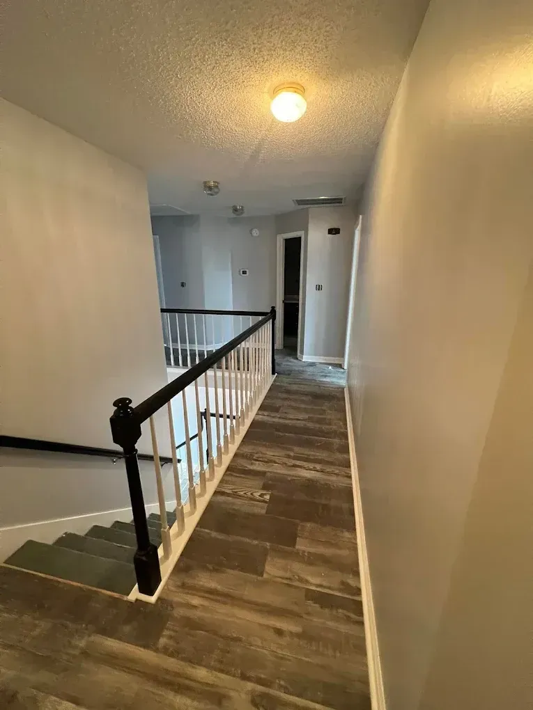 Narrow upstairs hallway with wood flooring, black railing, white walls, and a ceiling light.