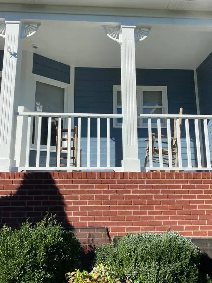 Blue front porch with white columns and railing above a brick foundation, with shrubs in front