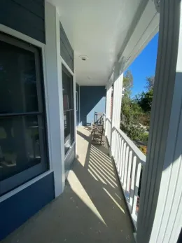 Sunny porch with white railing, blue walls, and rocking chairs overlooking greenery.