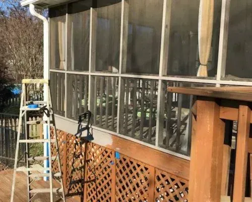 Screened porch with ladder, lattice skirting, and wooden deck railing in sunlight