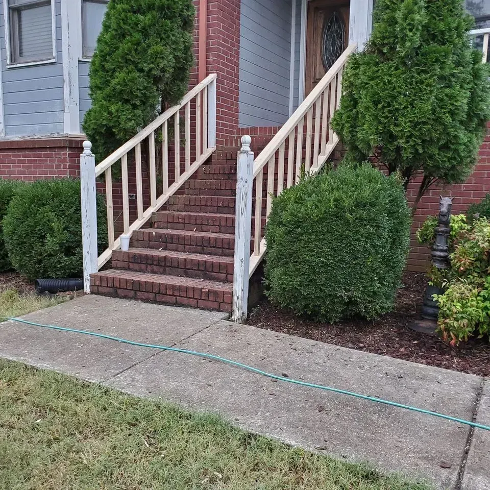 Front steps to a brick house with white railings, bordered by trimmed shrubs and a sidewalk.