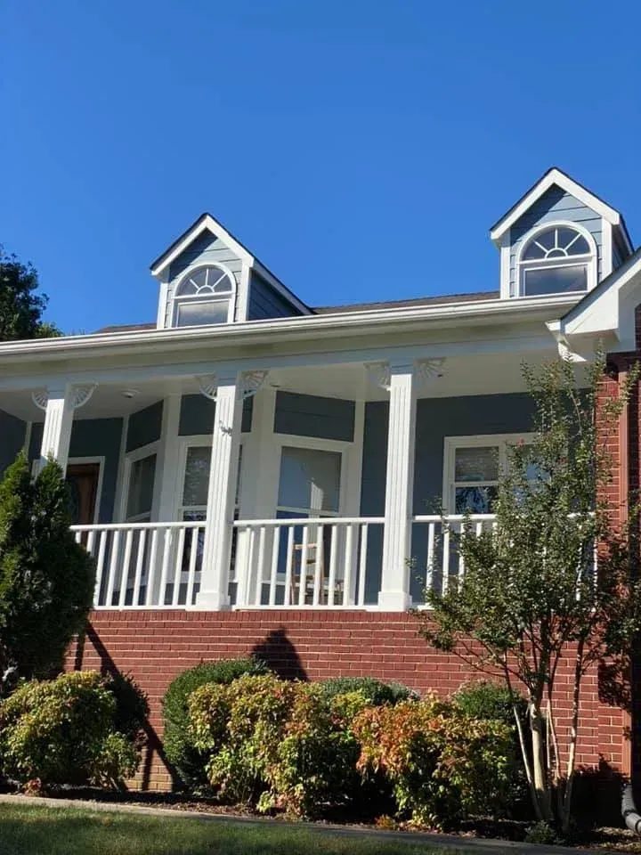 White house with red brick lower level, front porch, and dormer windows under a clear blue sky