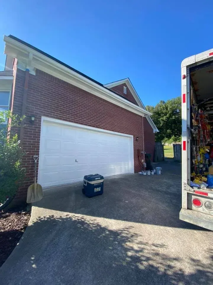 Brick house with a white garage door, moving truck at right, and bins on the driveway under a clear blue sky