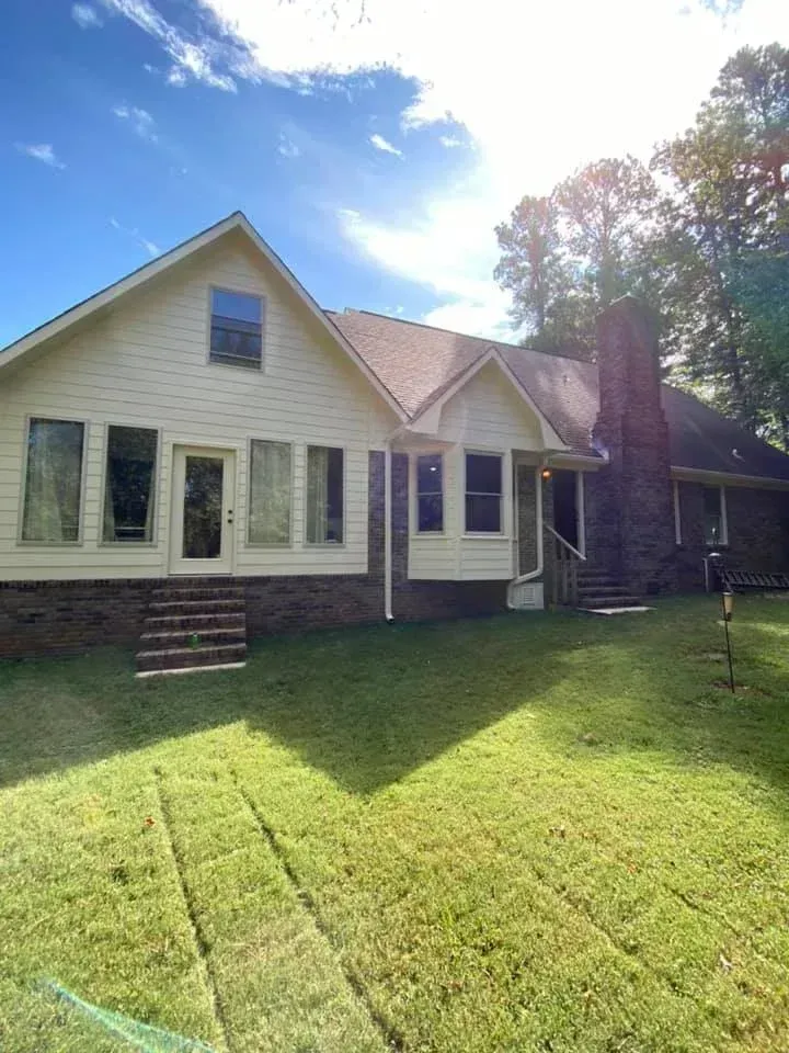 Sunlit beige house with brick foundation, front lawn, and tall trees under a blue sky