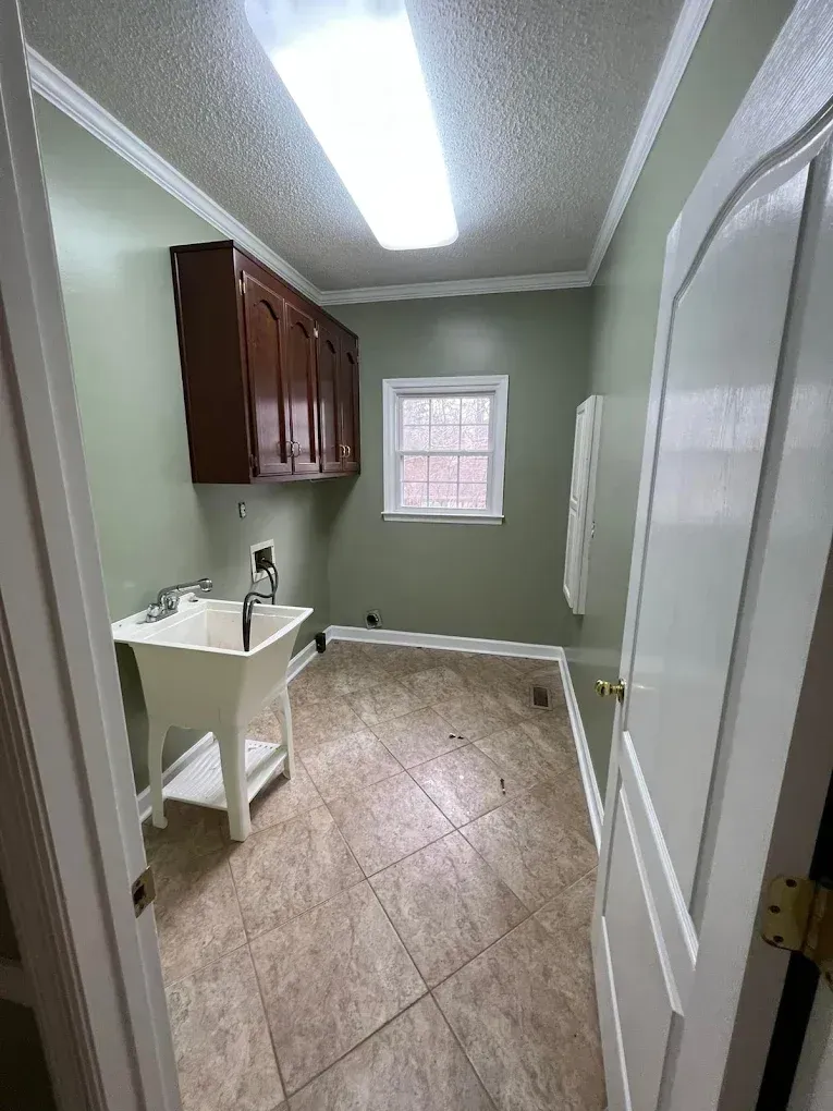 Small laundry room with sink, cabinets, window, and tiled floor under fluorescent light