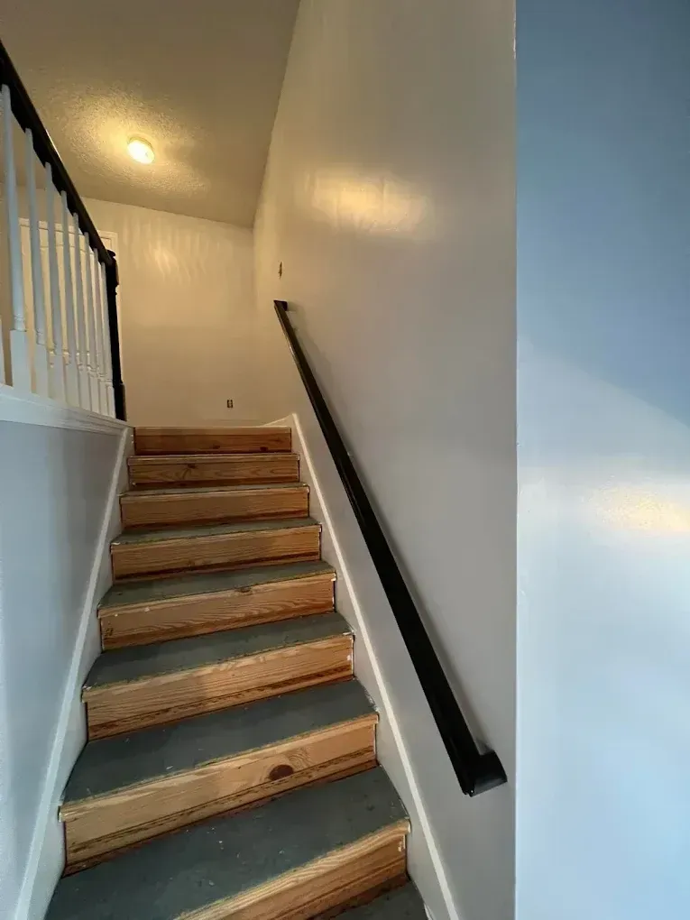 Staircase under construction with unfinished wooden steps, white walls, and a black handrail in a narrow hallway