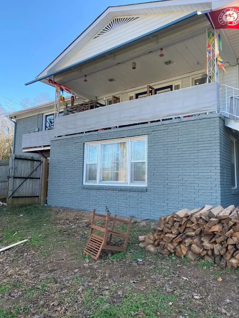 Two-story house with a covered balcony, gray brick lower level, and a stacked woodpile in the yard