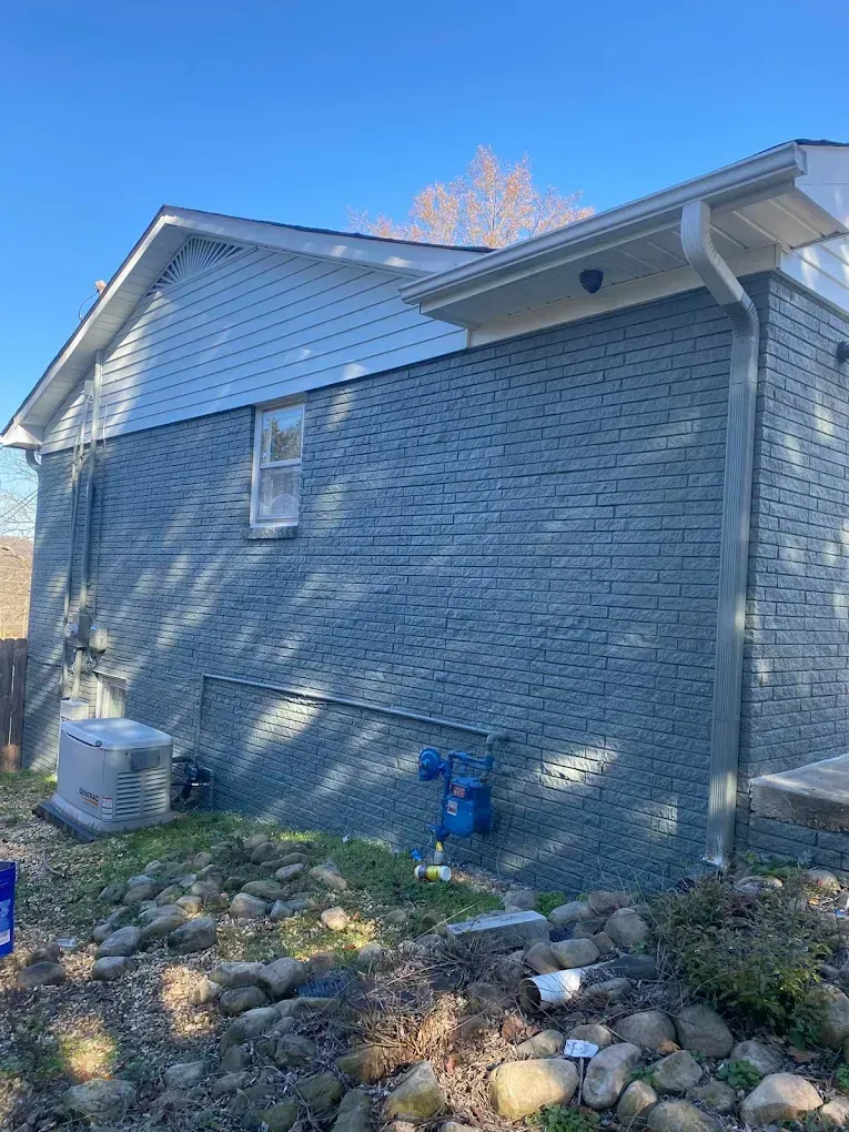 Side of a gray brick house with white siding, HVAC unit, and a rock-covered yard in sunlight