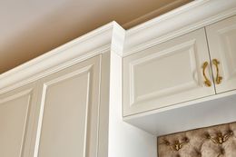 White cabinets with gold handles and crown molding above a tufted beige headboard