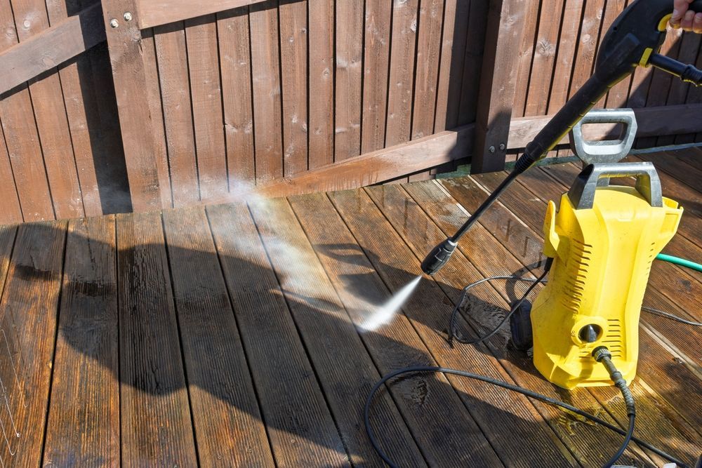 Yellow pressure washer cleaning a wooden deck beside a fence, spraying water onto the boards