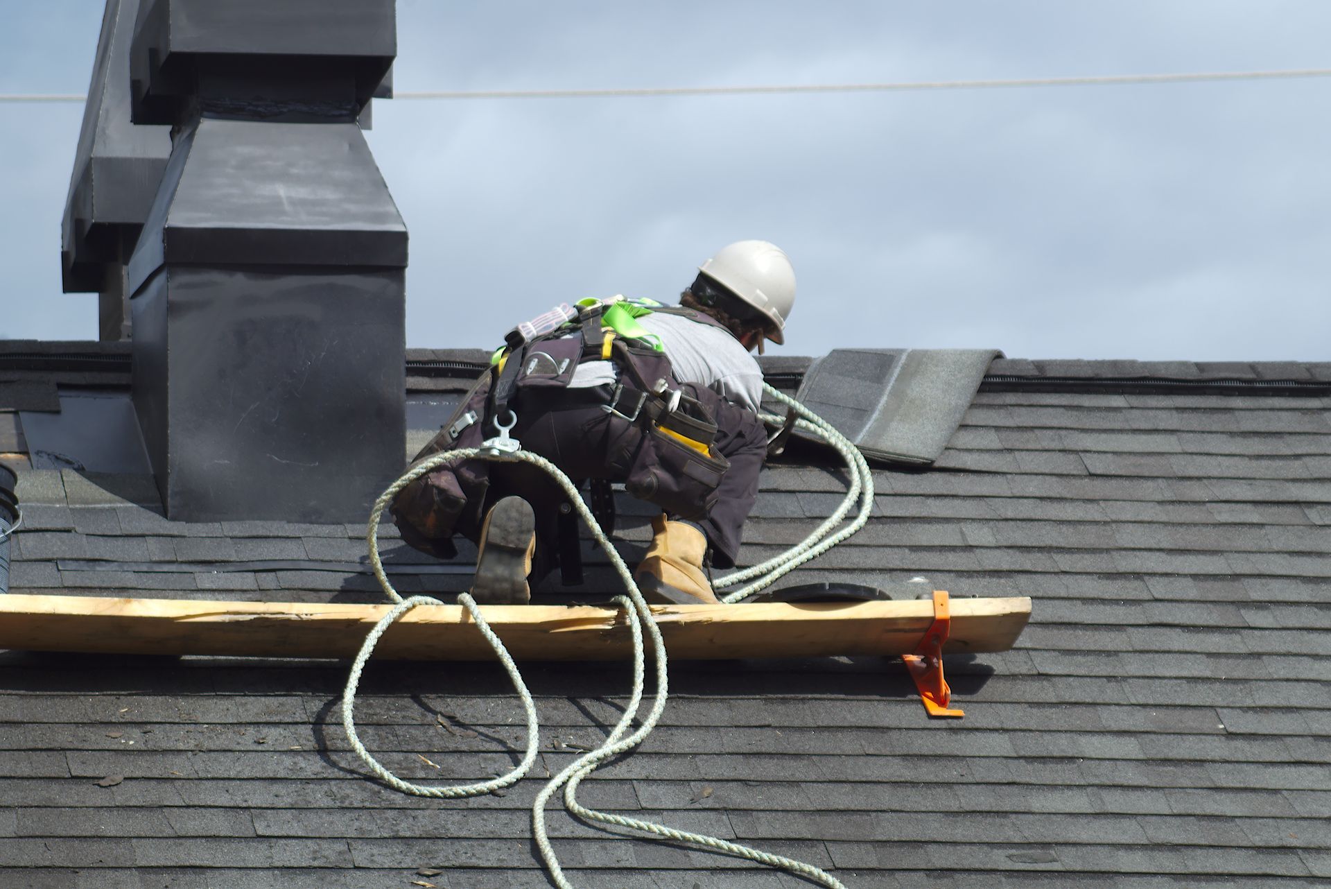 Worker on a shingled roof securing equipment near a chimney with hoses and tools visible