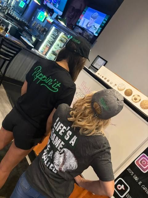 Two people in black shirts standing at a neon-lit bar counter, seen from behind.