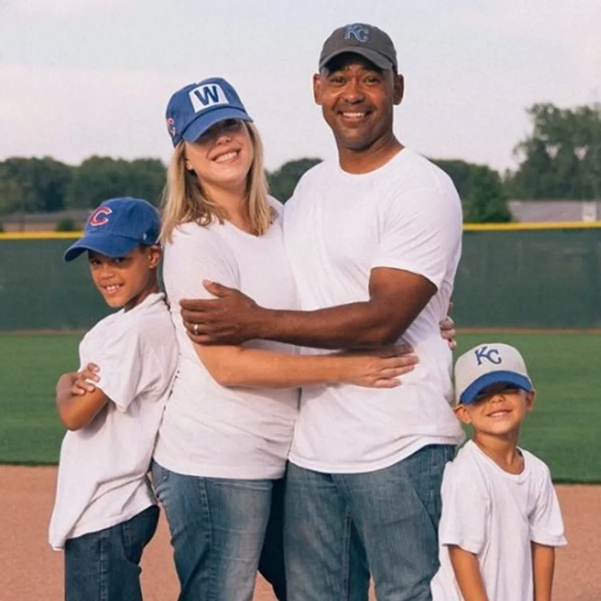 Family of four in white shirts and baseball caps smiling on a grassy field at dusk.