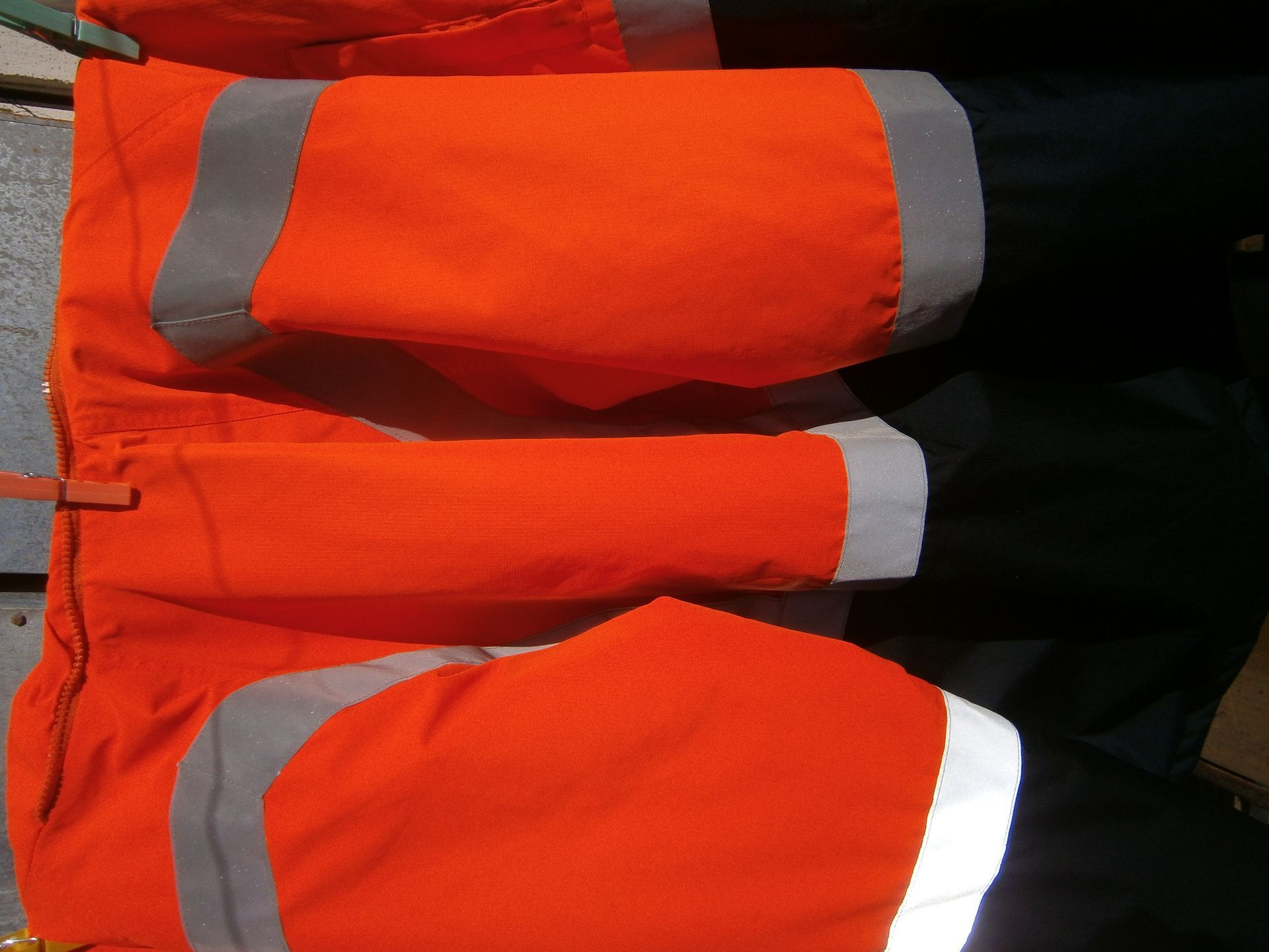 Pantalones de trabajo naranjas de alta visibilidad con tiras reflectantes grises, colgados en un tendedero al aire libre.