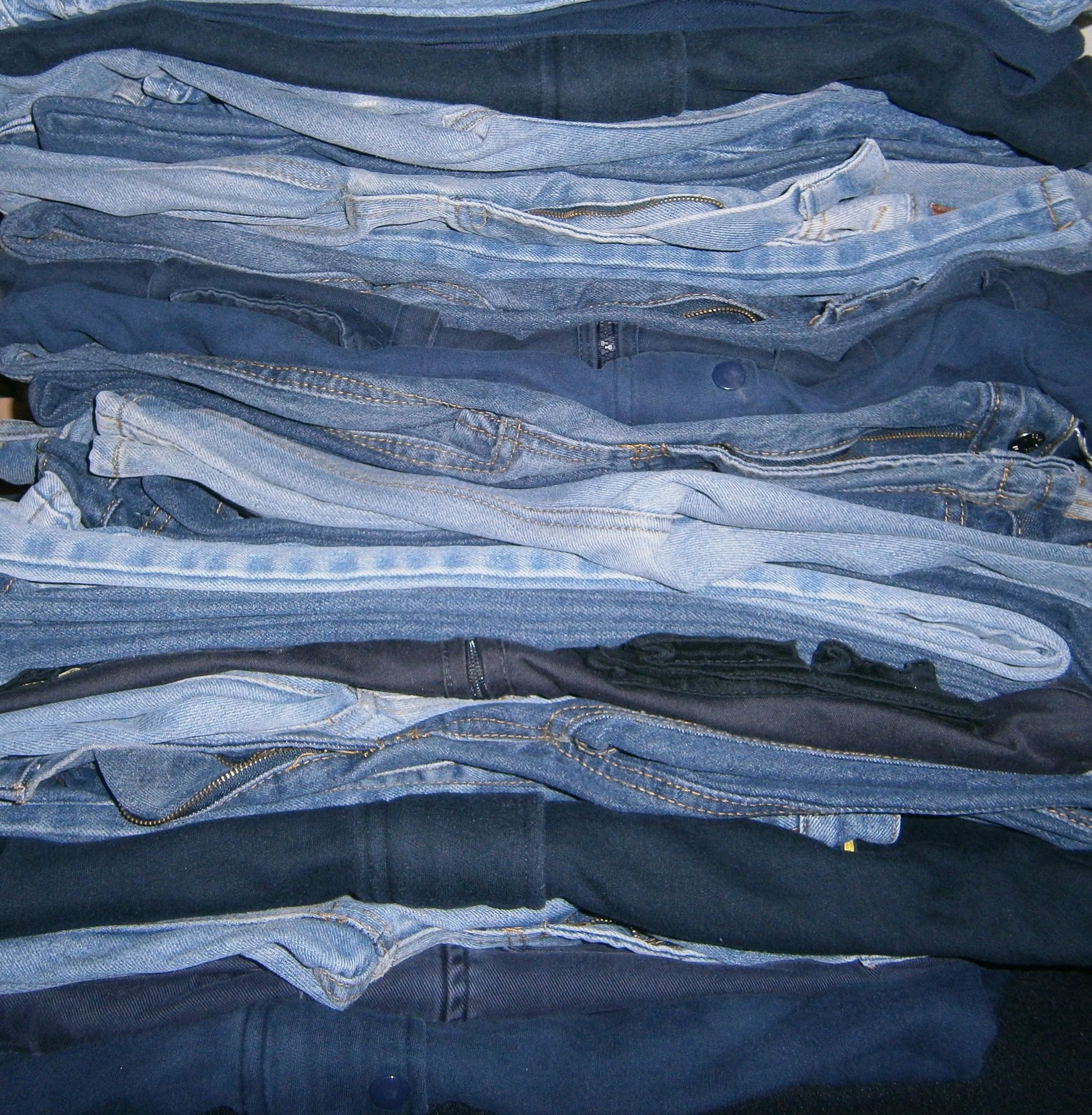 Una pila ordenada y vertical de pantalones vaqueros doblados y pantalones de colores oscuros en tonos azules y negros.