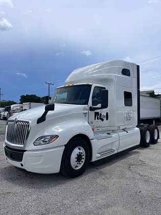 White semi-truck with trailer on an asphalt lot against a partly cloudy sky. The truck has the logo 