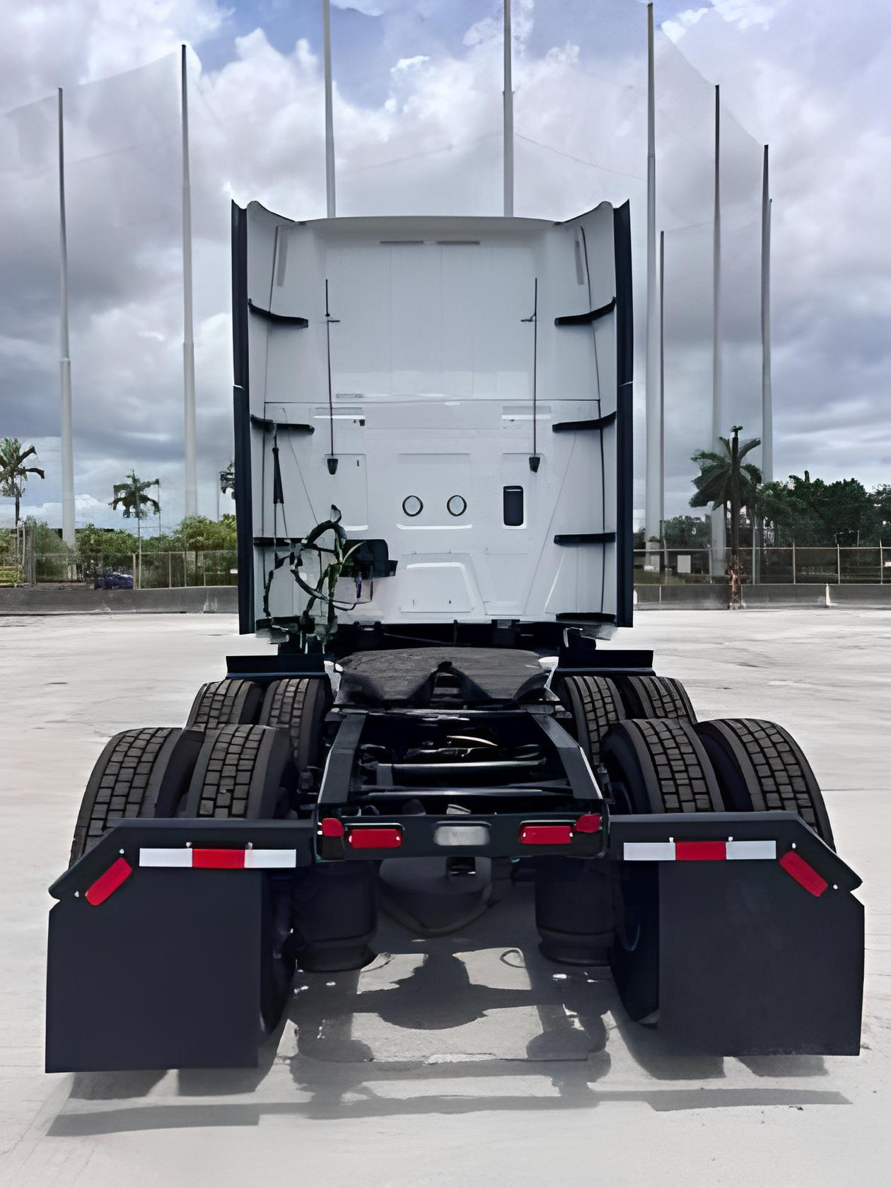 Rear view of a white semi-truck cab and chassis with black fenders and tires, against a cloudy sky.
