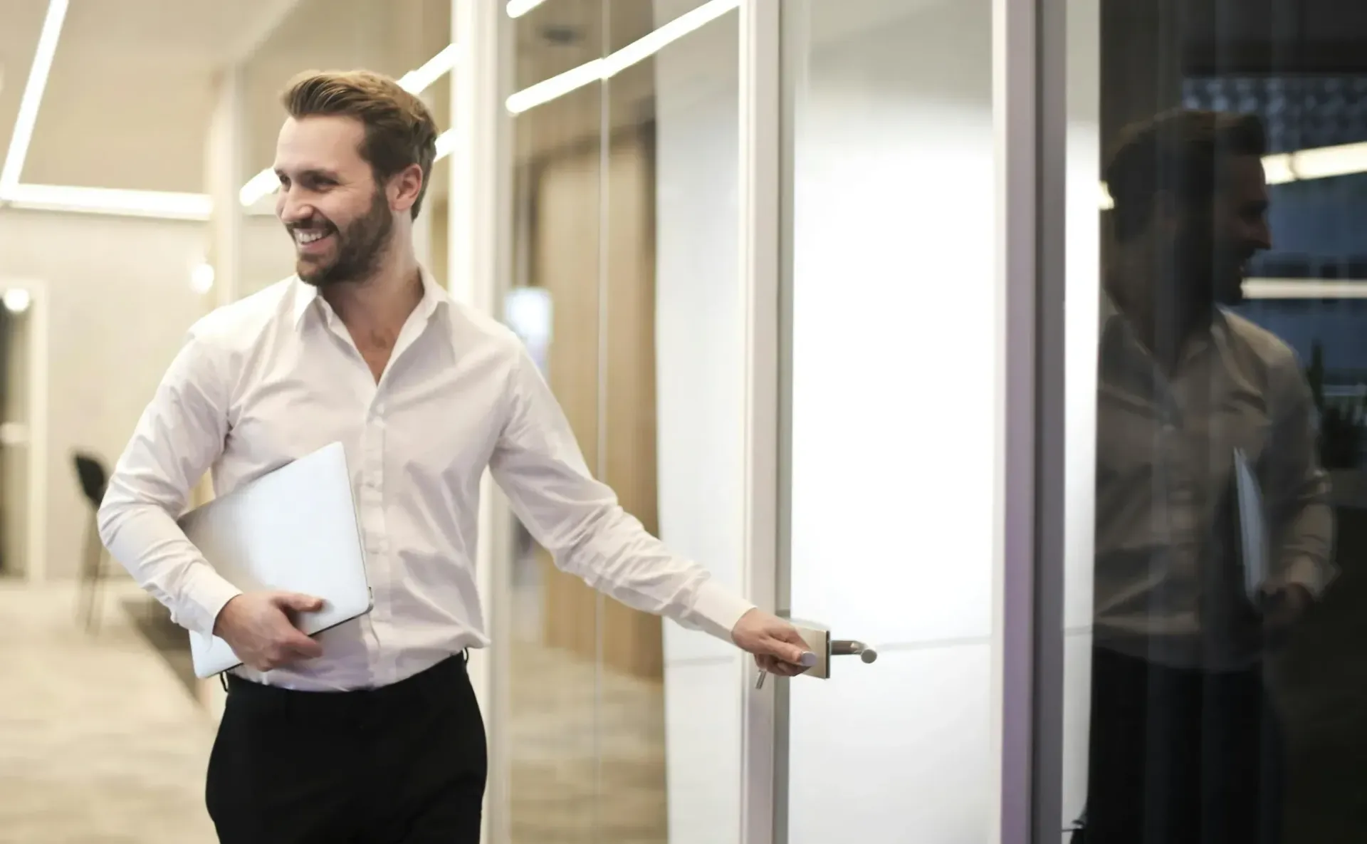 A man is holding a laptop and opening a door in an office.