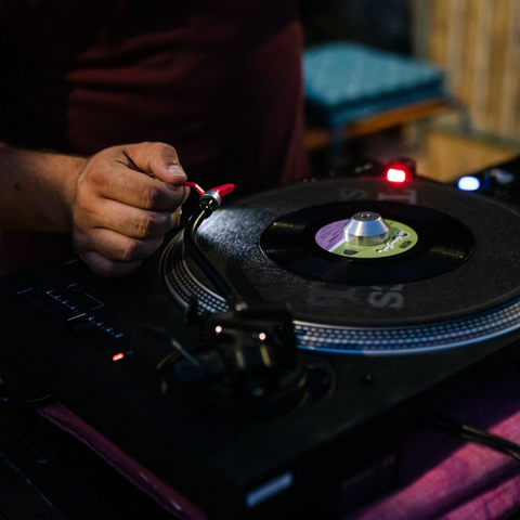 In un ambiente scarsamente illuminato, una mano manovra il braccio di un giradischi nero che riproduce un disco in vinile.