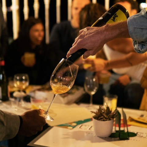 In un ambiente scarsamente illuminato, una persona versa del vino in un bicchiere tenuto da un'altra.