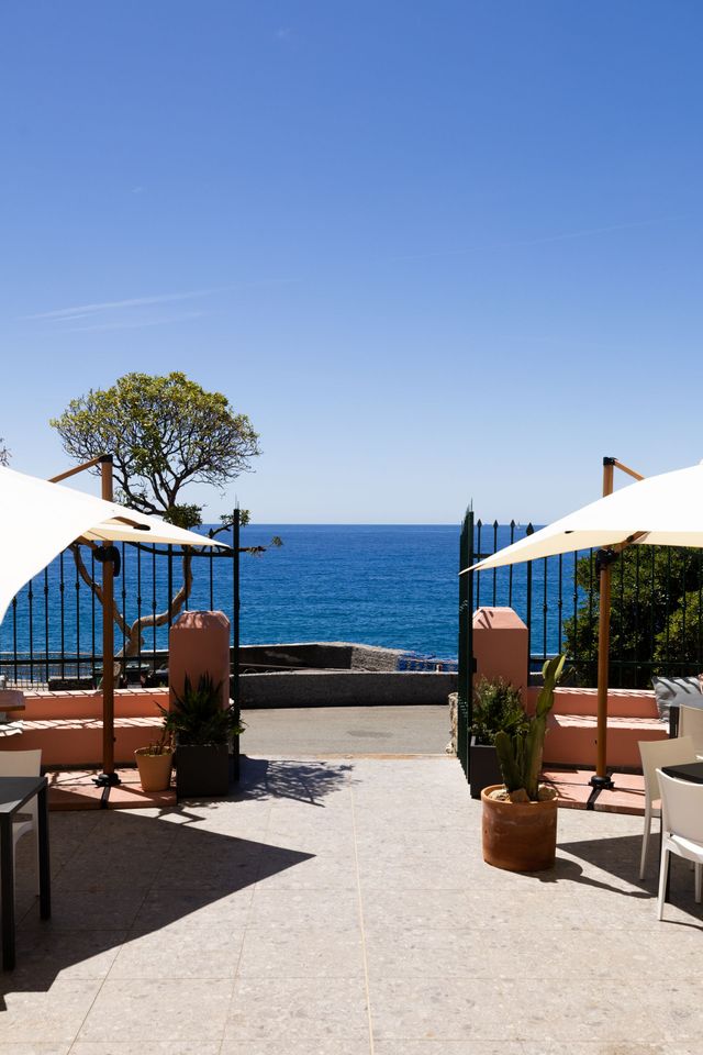 Patio con vista sull'oceano, ombrelloni bianchi, piante in vaso e un cielo azzurro limpido.