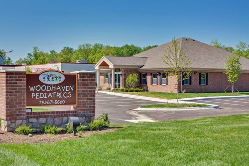 Woodhaven Pediatrics brick office building with sign, driveway, and landscaped lawn on a sunny day