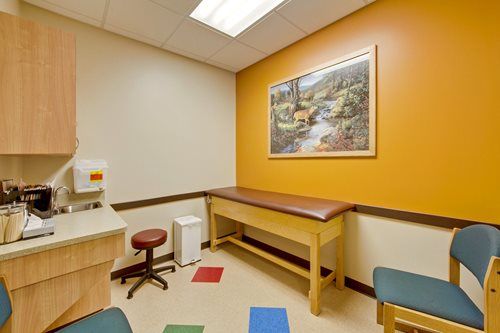 Exam room with wooden exam table, stools, sink, and a landscape painting on an orange wall.