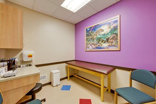 Small medical exam room with purple accent wall, exam table, chairs, and a landscape painting.