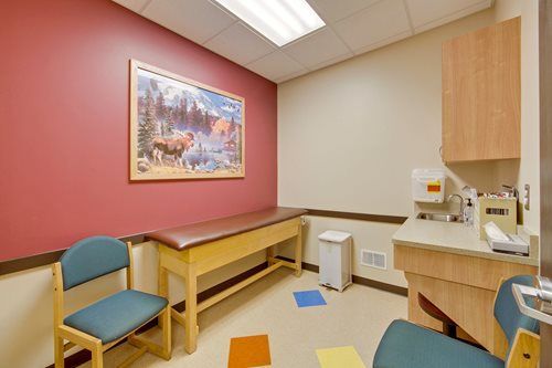 Empty medical exam room with chairs, exam table, sink, and wall painting in soft pink and beige tones