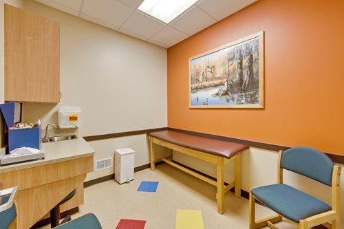 Exam room with exam table, sink, cabinets, and two blue chairs against orange and beige walls