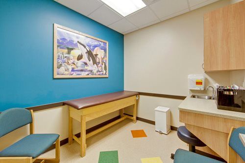 Small exam room with teal walls, exam table, chairs, cabinets, and a coffee maker.