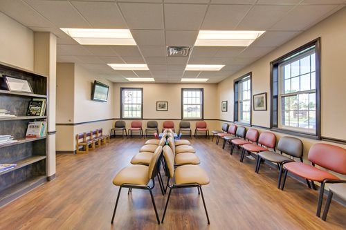 Empty waiting room with rows of chairs, wood floor, and large windows letting in daylight
