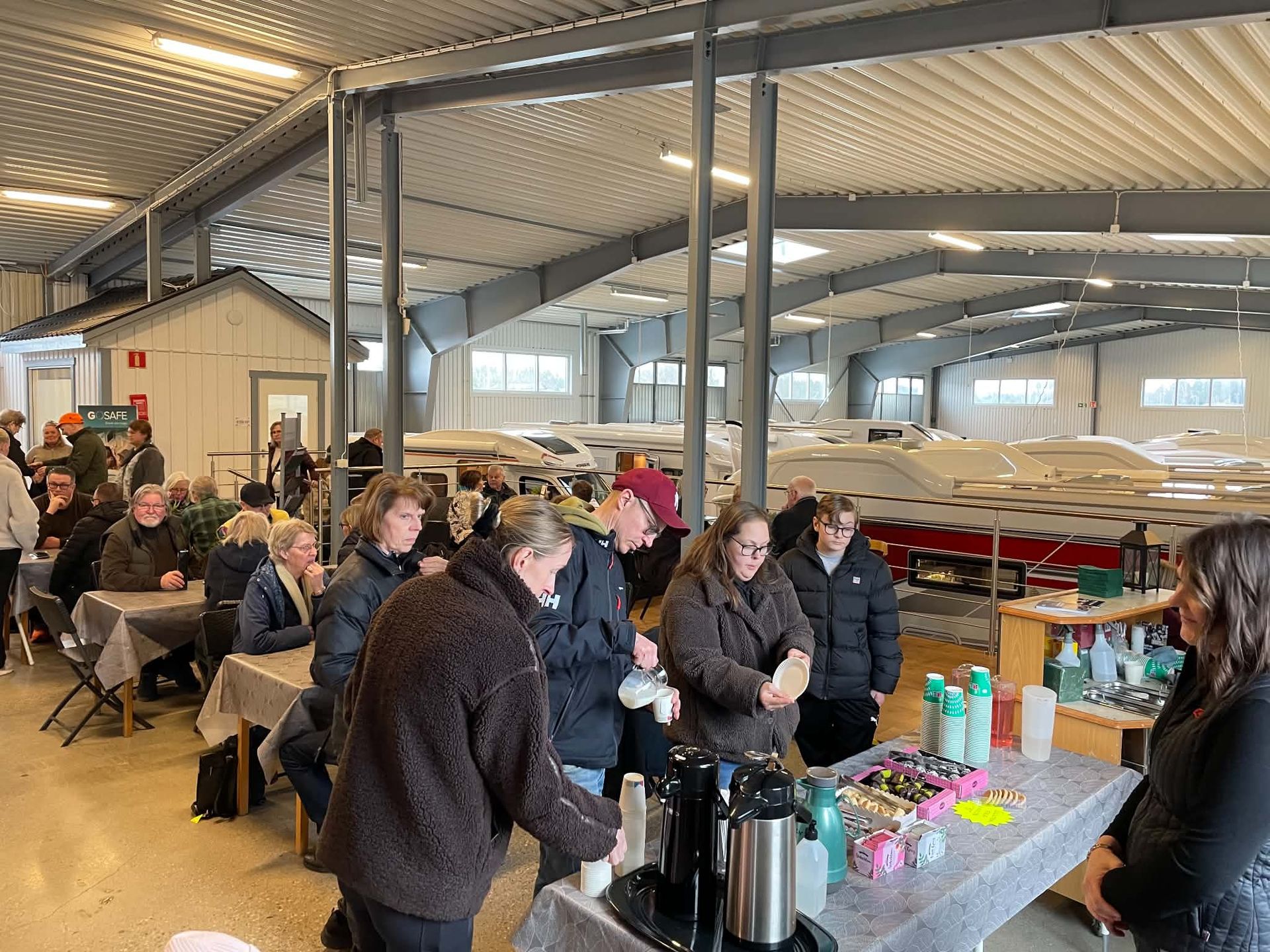 People gathered in a bright indoor hall around a refreshment table with coffee and drinks.