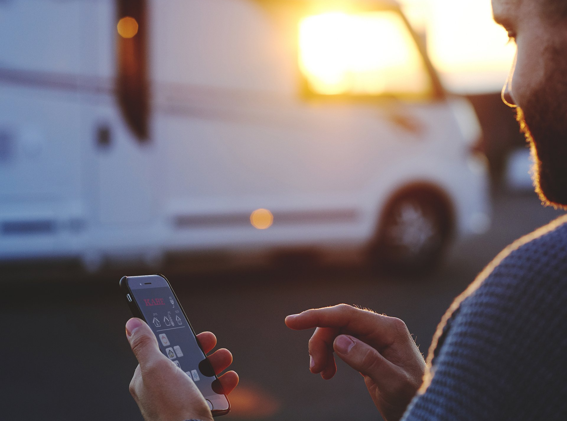 Man using a smartphone with a recreational vehicle in the background, likely controlling RV functions at sunset.
