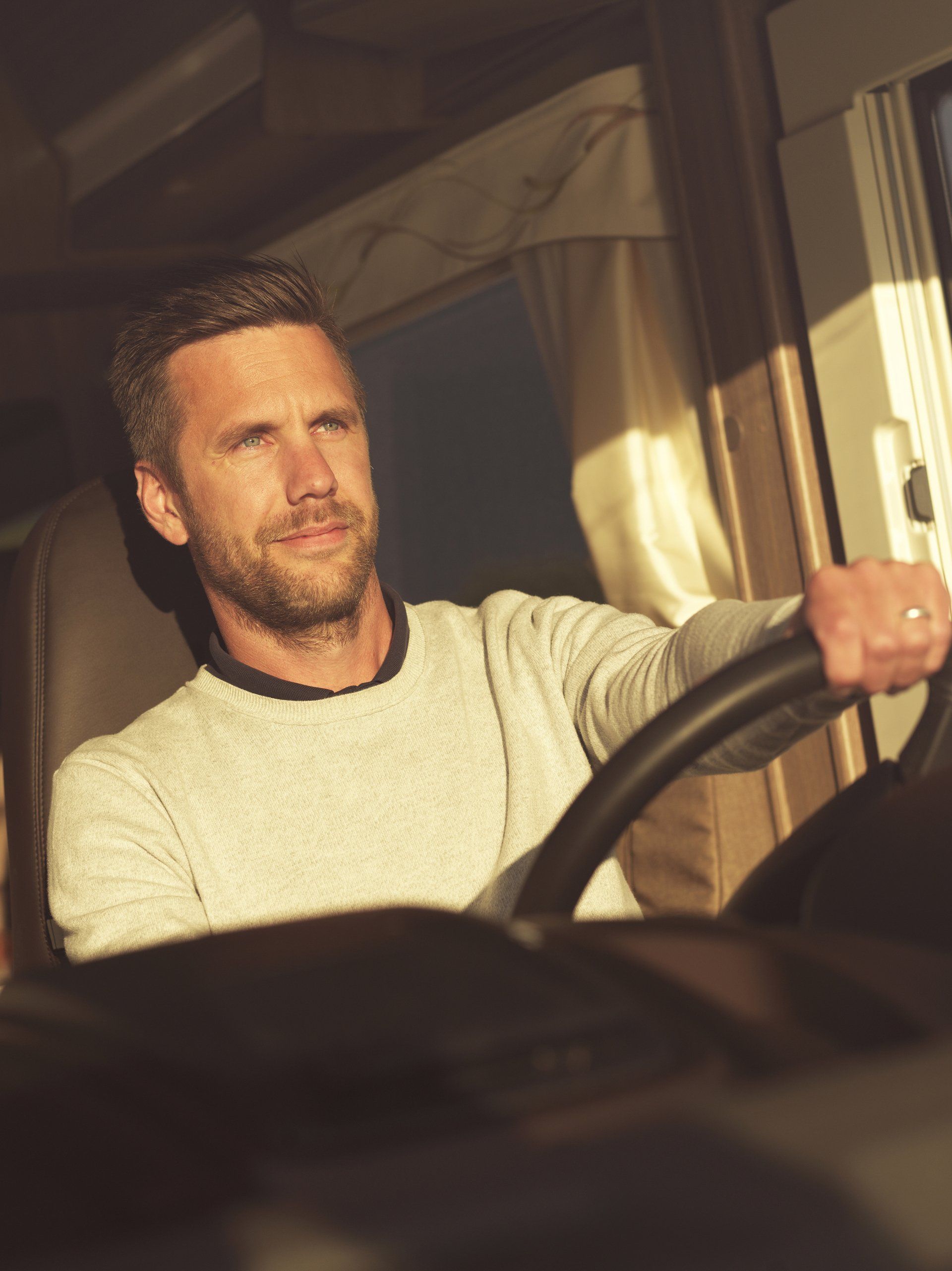 Man driving a vehicle, looking to the side. Wearing a light sweater. Sunlight streams in.