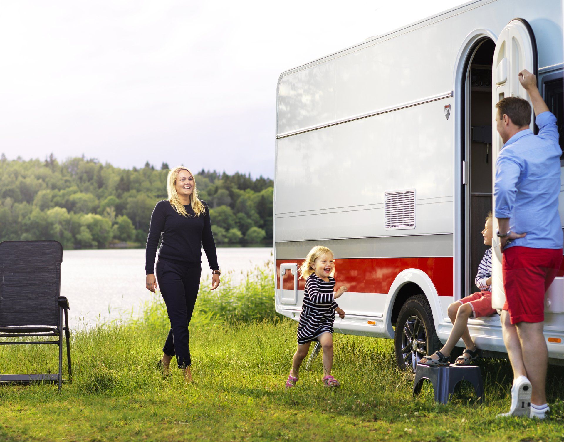 Family near a white and red camper by a lake; woman walks toward it, man by the door.