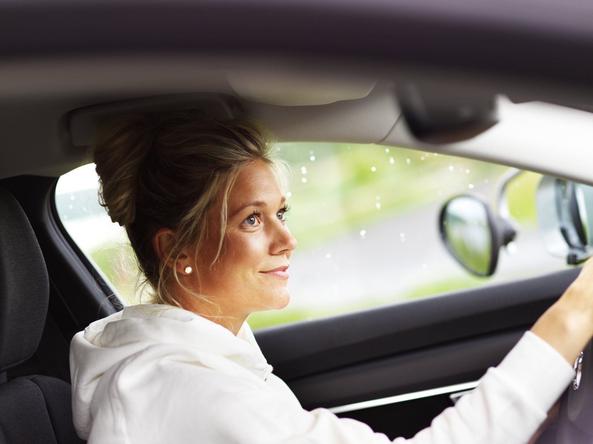 Woman driving a car, looking ahead with a side view mirror visible, wearing a white hoodie.