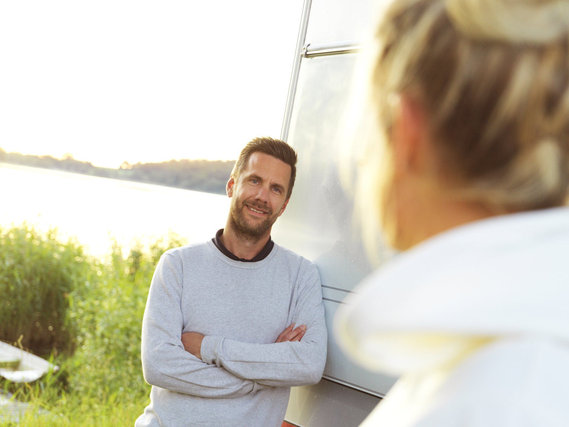 Man with arms crossed, smiling, leaning against a silver camper, looking at a person with their back to the camera near a body of water.