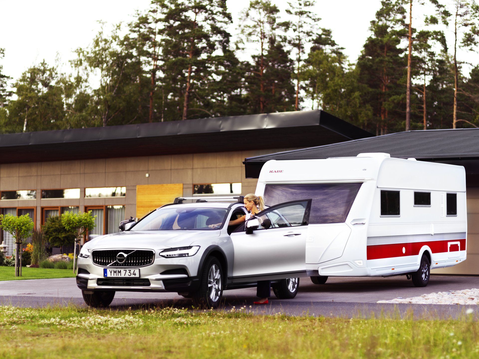 Silver car towing a white and red camper in front of a modern house. Person exiting car.