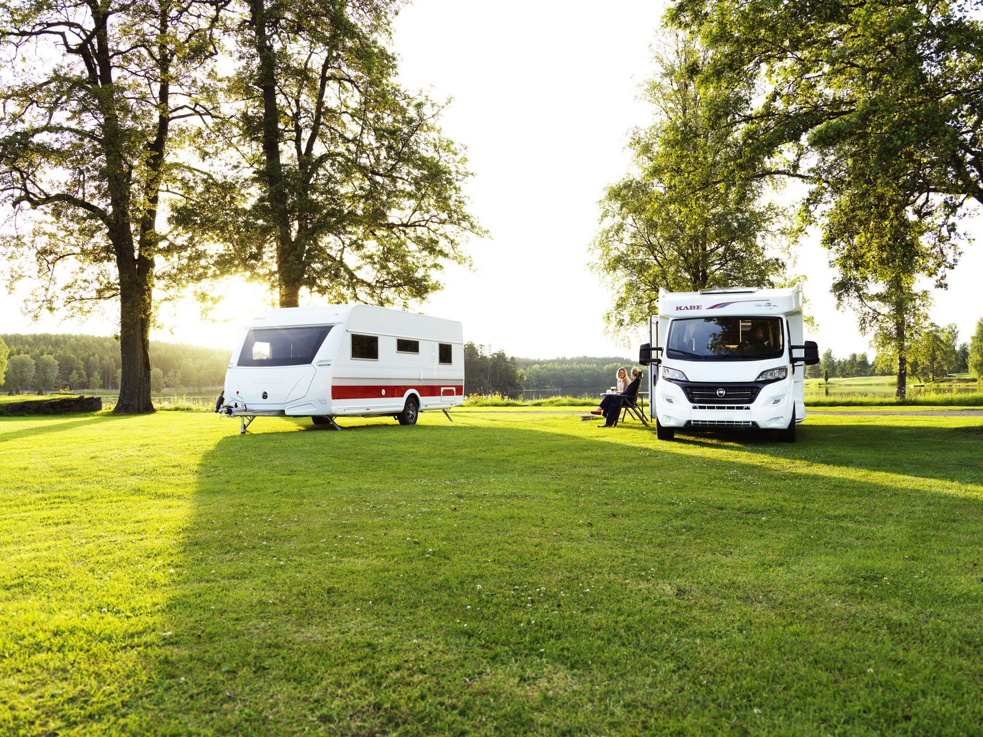 Caravan and camper van on a grassy field with trees. Person sitting near the camper.