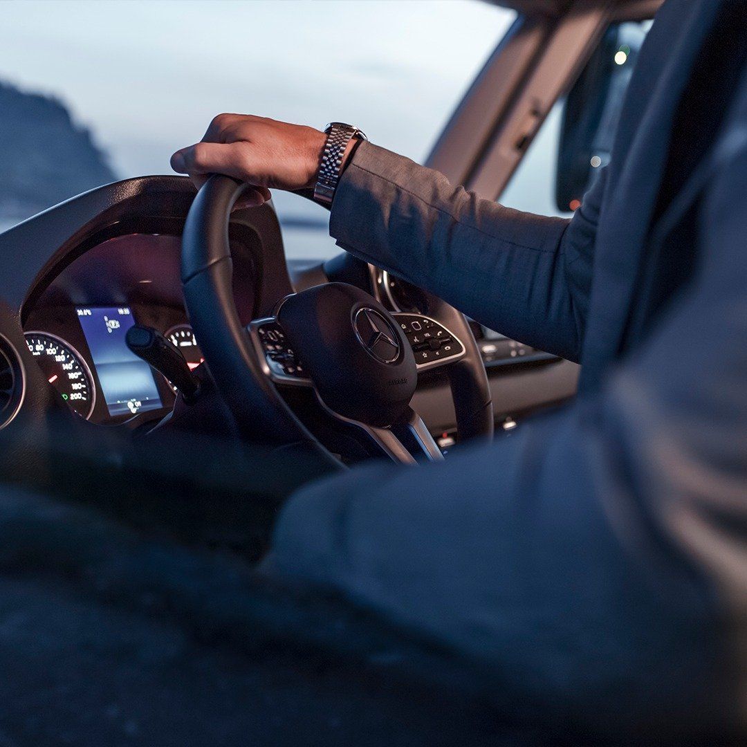 Person driving a vehicle, hands on steering wheel. Dark interior, visible dashboard, and a view of the outside.