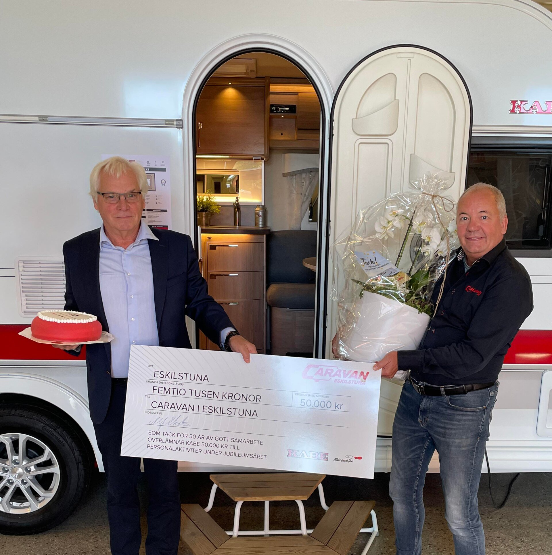 Two men in front of a camper. One holds a check, the other, a cake and flowers.