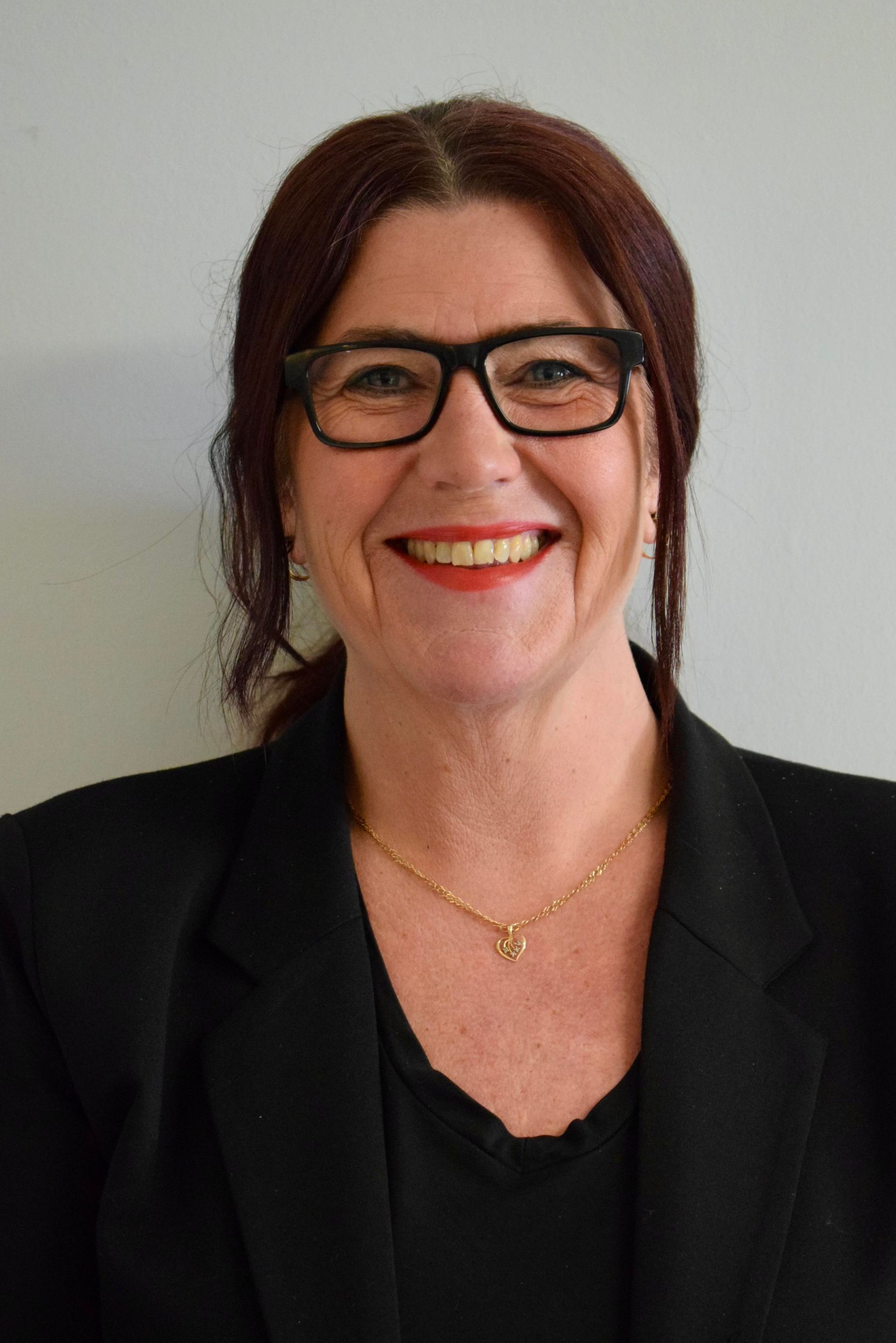 Woman with glasses and red lipstick, wearing black blazer, smiling.