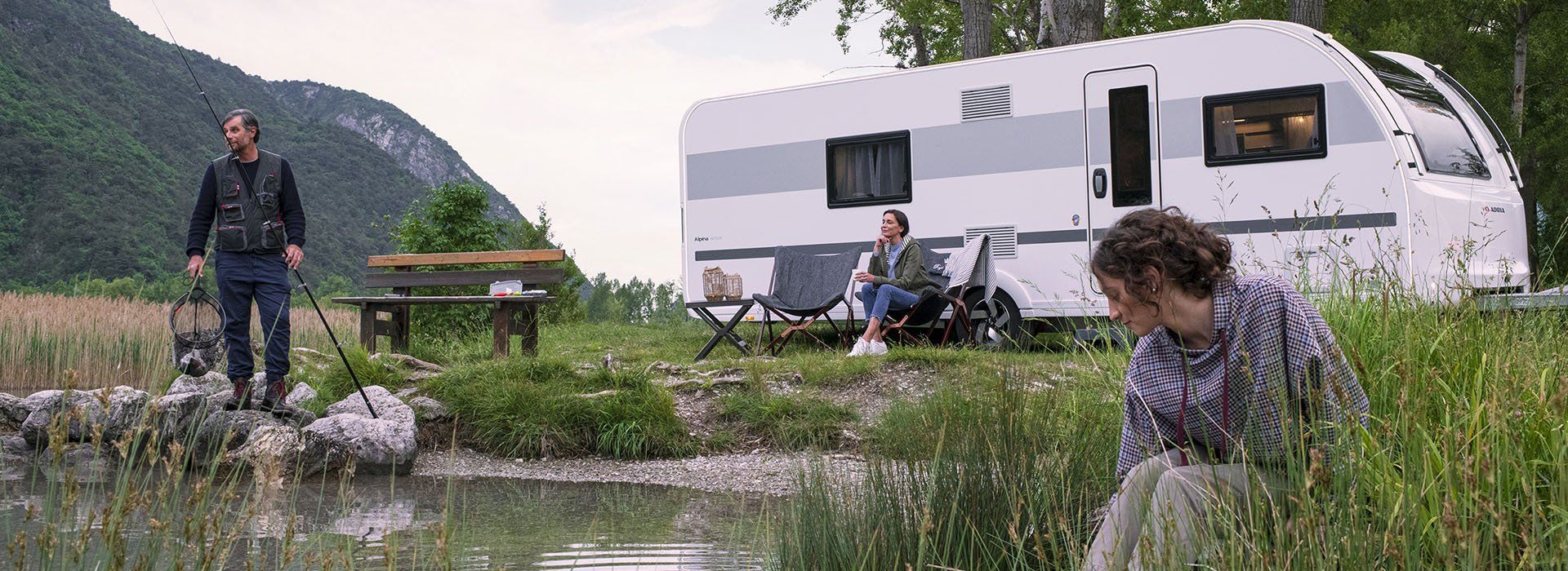 A man fishes by a lake near a campsite with a camper and two people sitting outside.
