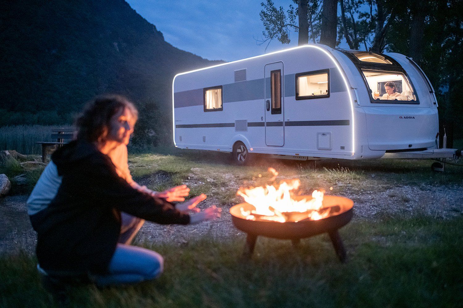 Two people by a campfire near a lit white RV at dusk, surrounded by trees and a mountain.